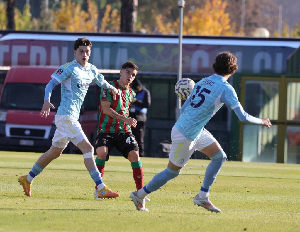 Two young football players in training, one wearing a blue jersey with 'Juventus' branding and the other in a striped red and green jersey, as they compete for the ball on a grassy field.