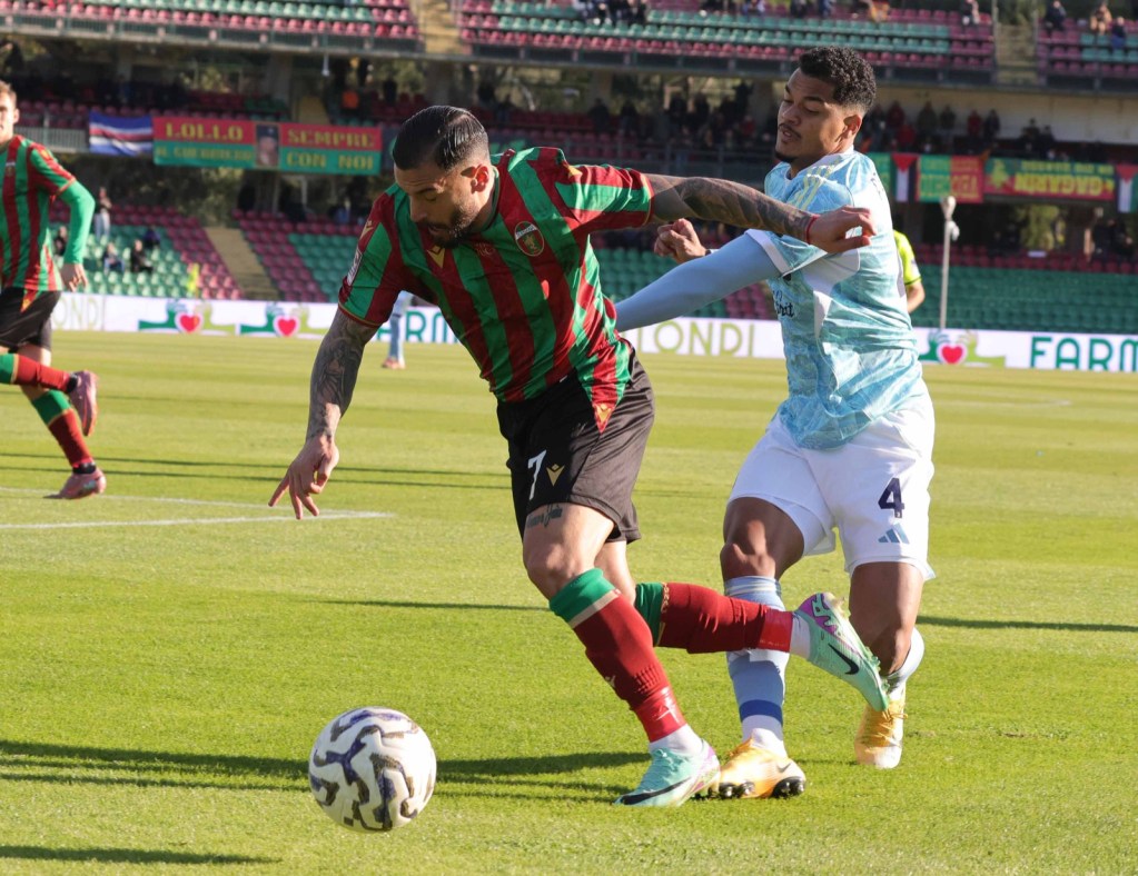 Two football players competing for the ball on a grassy field during a match. One player is wearing a green and red striped jersey while the other is in a light blue jersey.