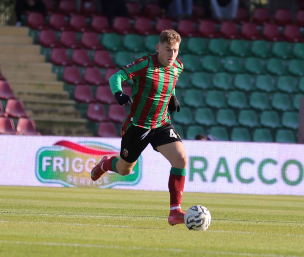 A soccer player in a striped green and red jersey sprinting on the field, with a soccer ball at his feet and a blurred background of stadium seats.