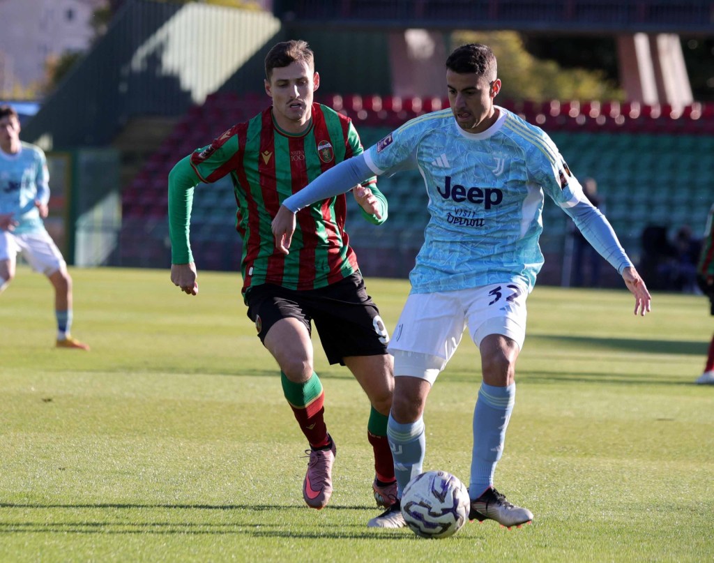 Two soccer players competing for the ball on a grassy field. One player is wearing a green and red striped jersey while the other is in a light blue jersey.