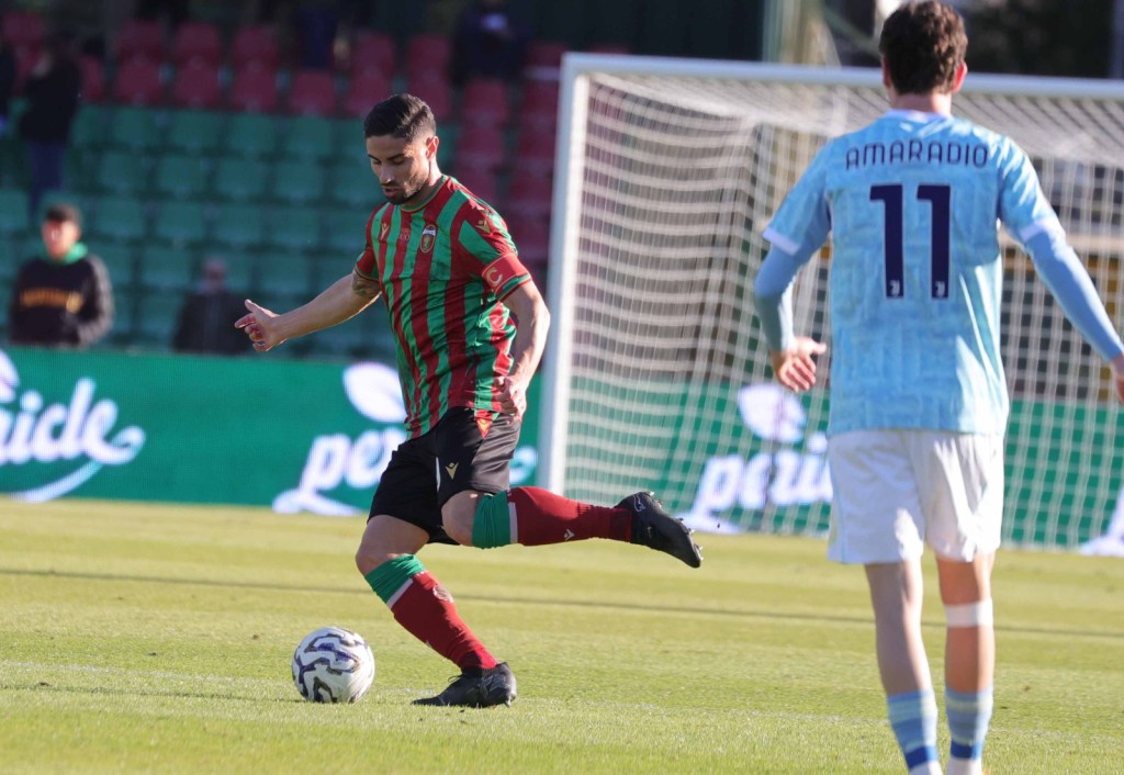 A football player in a red and green striped jersey is kicking the ball on the field, while another player in a light blue jersey (number 11) stands nearby.