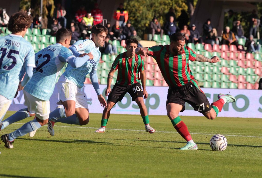 A soccer player wearing a red and green jersey kicks the ball while being challenged by defenders in light blue jerseys on a grassy field.
