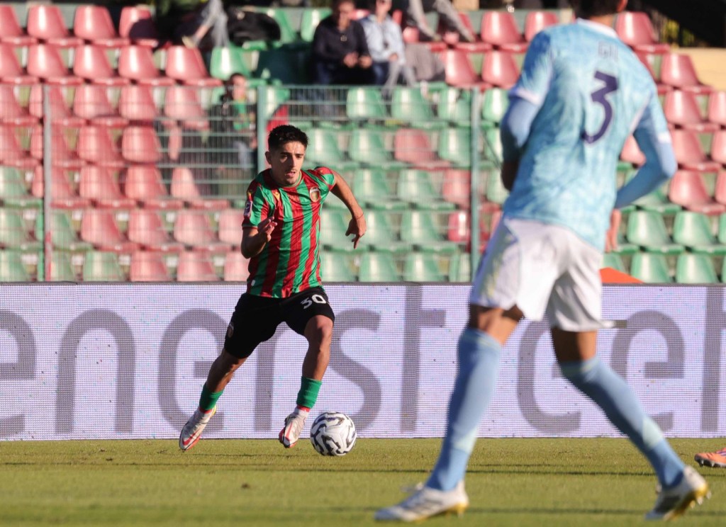 A soccer player wearing a green and red striped jersey is sprinting on the field, while another player in a light blue jersey is watching. The background shows empty stadium seats.