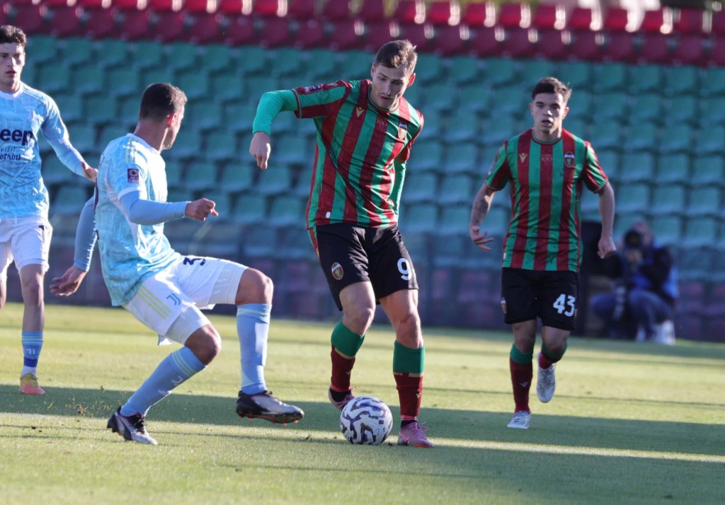 Football players in action during a match; one player in a red and green striped jersey dribbles the ball while two opponents in light blue jerseys attempt to defend.