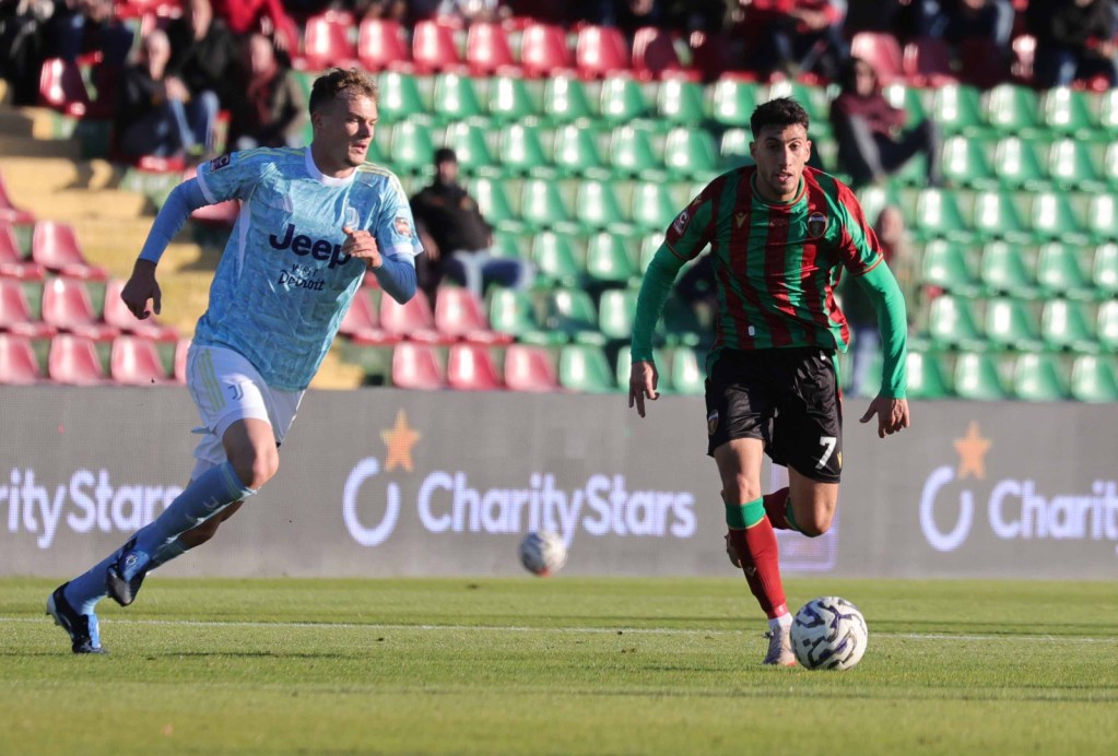 A soccer match in progress with two players sprinting towards the ball. One player is wearing a light blue jersey with 'Jeep' branding, while the other is in a green and red striped jersey. The background features a stadium with spectators.
