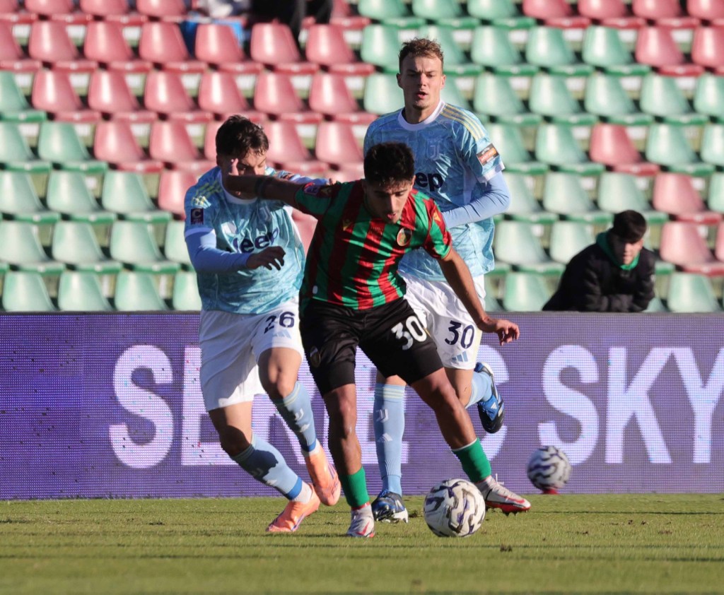 Two football players in action on the field, one in a red and green striped jersey and the other in a light blue jersey. A third player is behind them, wearing a different blue jersey.