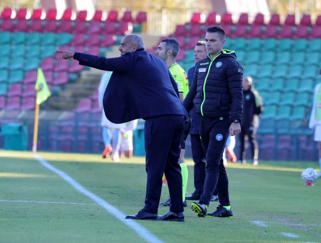 A football coach gestures from the sidelines during a match, while several team members and officials are visible in the background.
