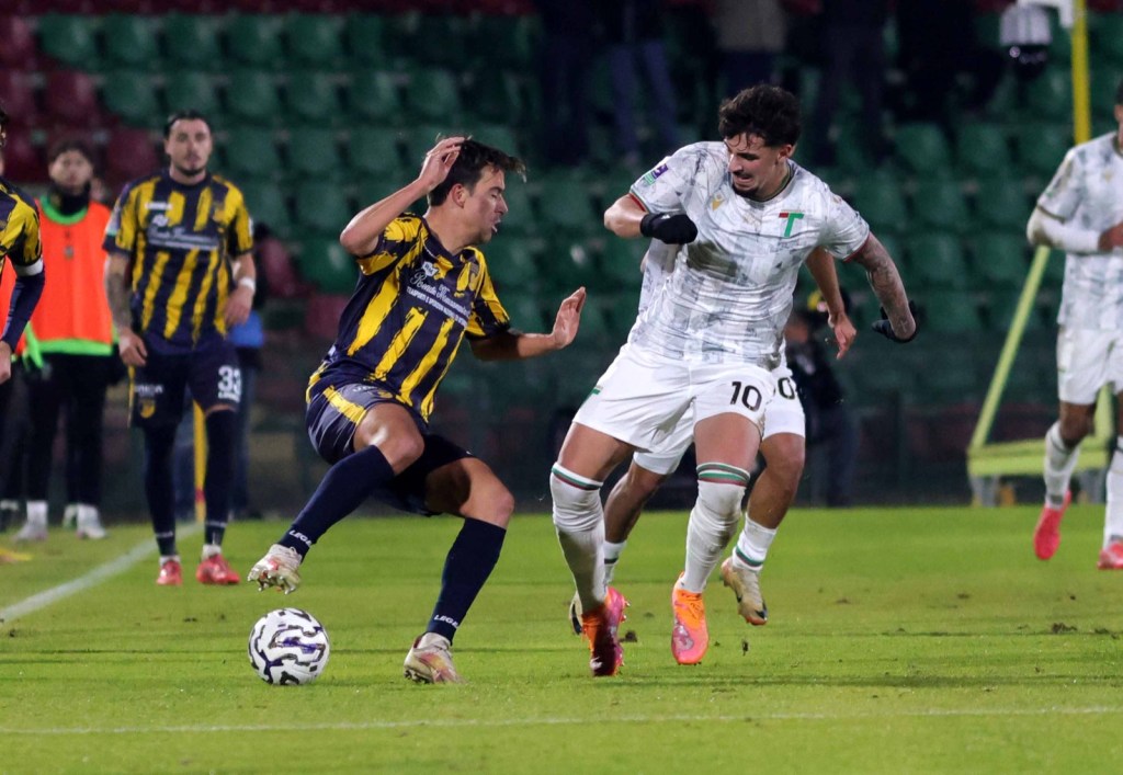 A tense moment during a soccer match with two players in action, one wearing a striped yellow and blue jersey and the other in a white and green kit. The player in yellow appears to be falling while the player in white is lunging towards him.