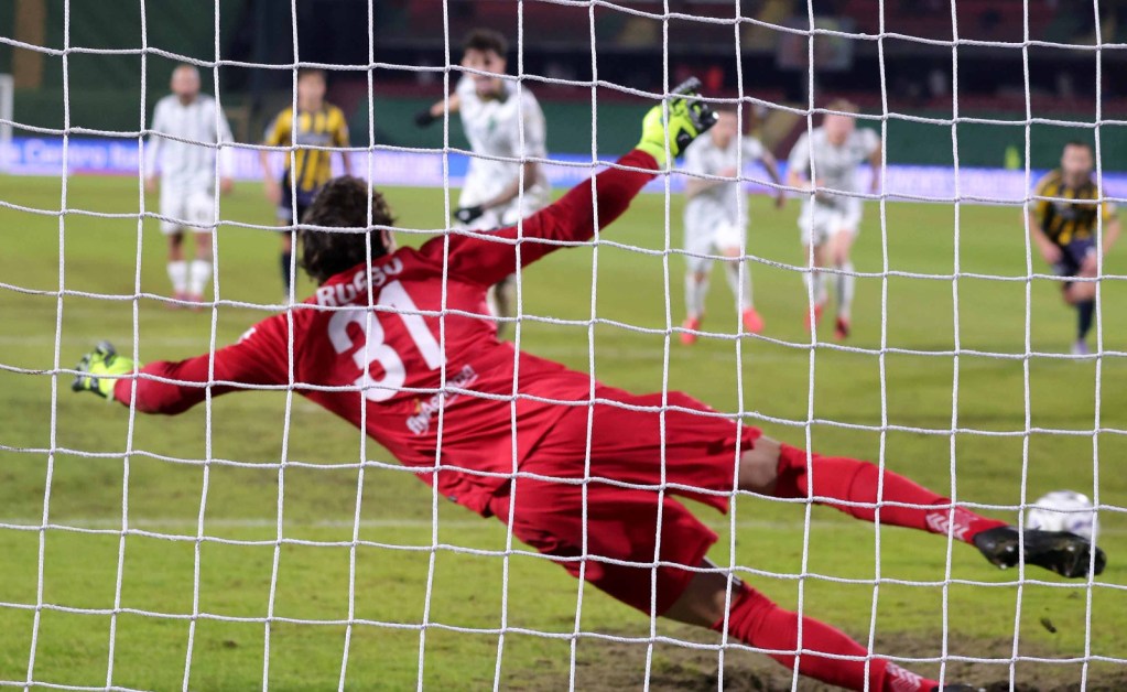 A goalkeeper in a red uniform dives to save a penalty kick while players stand in the background.