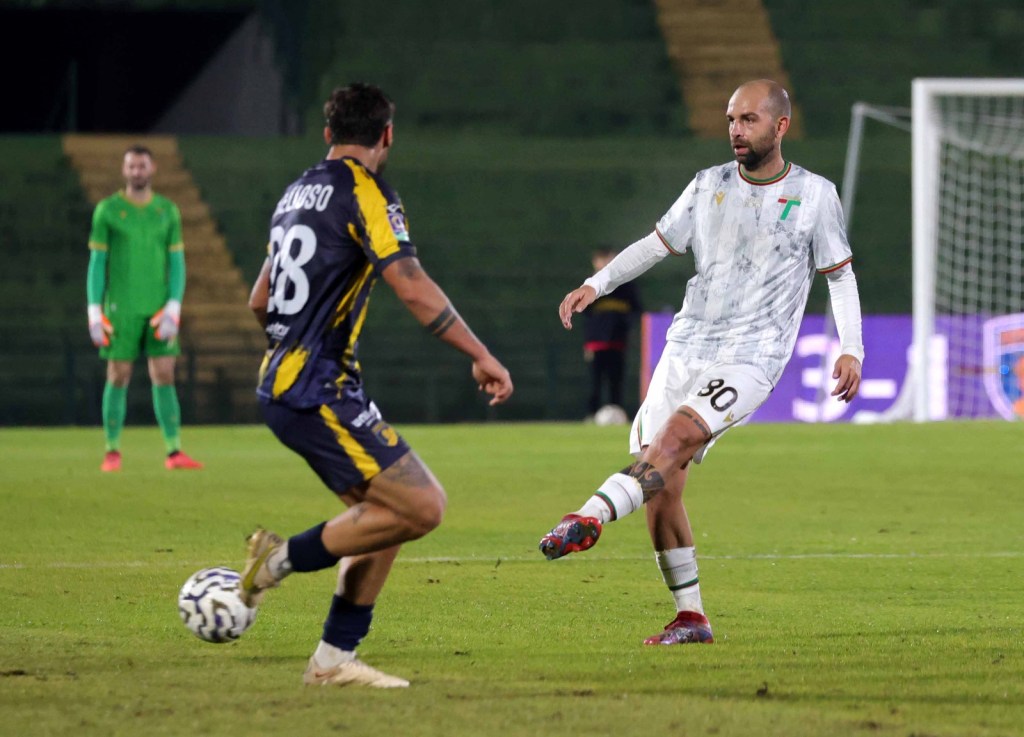 Un jugador de fútbol en acción, vestido con un uniforme blanco y verde, está pateando el balón mientras un oponente, con un uniforme oscuro y amarillo, lo observa. Al fondo, se puede ver al portero en uniforme verde.
