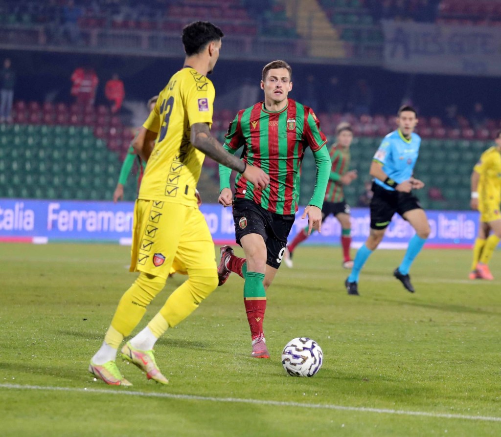 A soccer match in progress, featuring two players: one in a yellow goalkeeper uniform and the other in a red and green striped jersey, with a soccer ball at their feet.