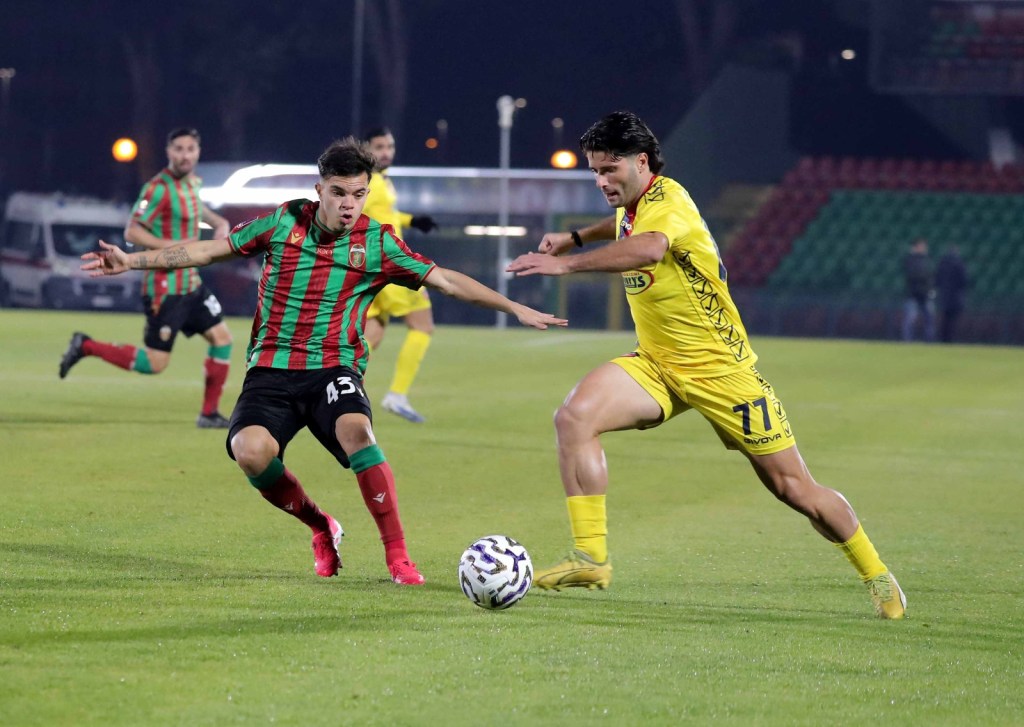 Two soccer players in action on a field, one in a red and green striped jersey and the other in a yellow jersey, competing for the ball under stadium lights.