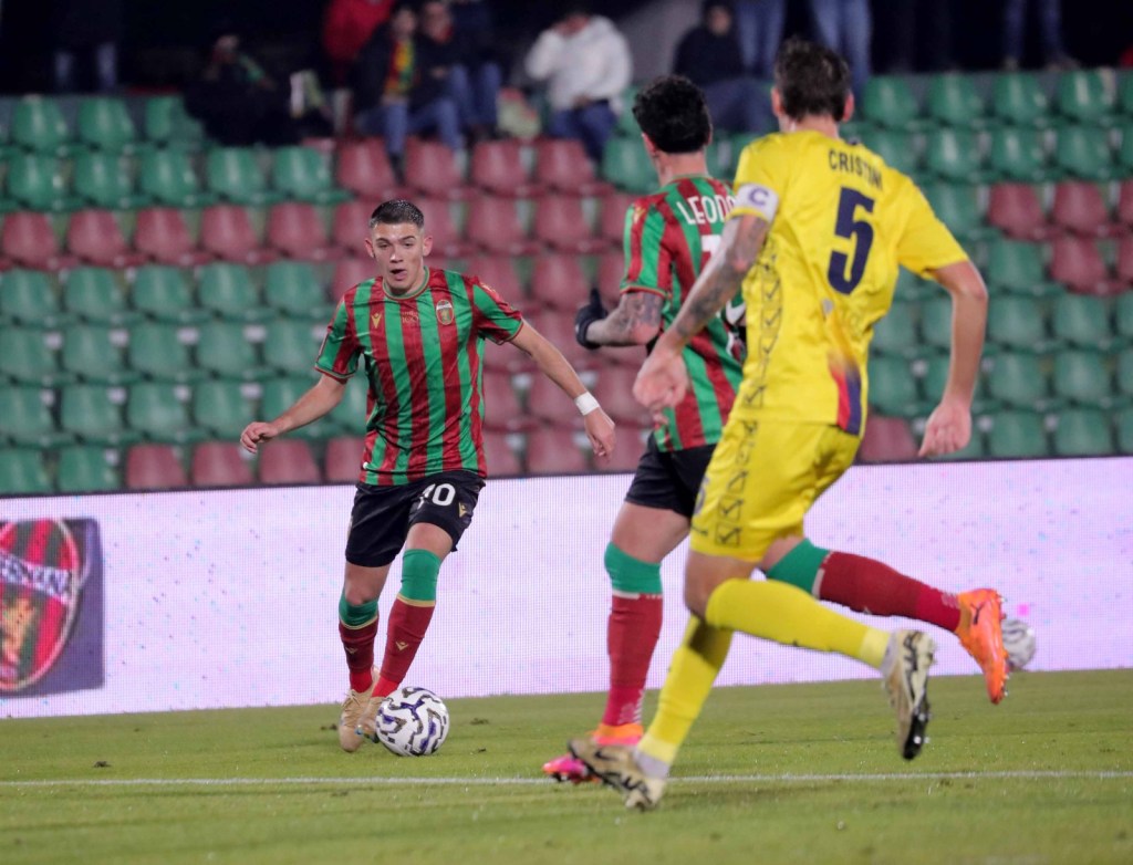 A football match in progress with players from two teams competing. One player in a red and green striped jersey dribbles the ball, while others are in action in the background, showcasing a vibrant stadium atmosphere.