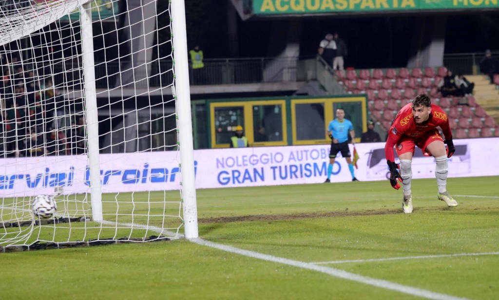 A goalkeeper in a red uniform is rushing towards the goal post as the ball crosses the goal line, indicating a goal scored during a soccer match.