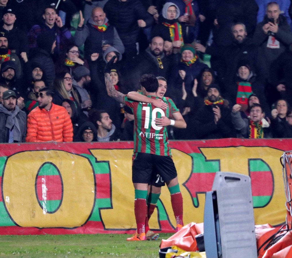 Two soccer players celebrating a goal, embracing each other on the field. In the background, enthusiastic fans are cheering with a colorful banner.