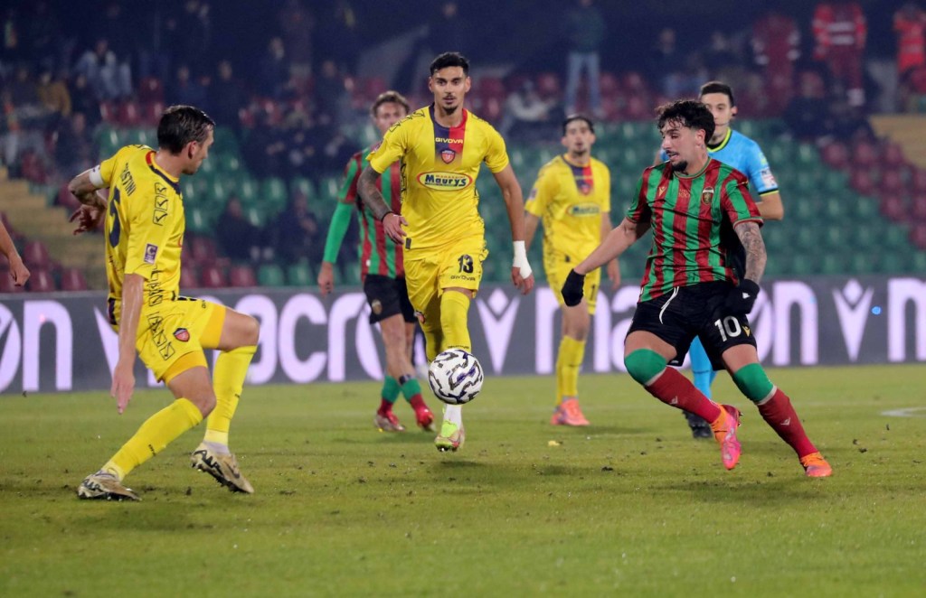 A soccer match in action, featuring players in yellow and green-red kits. One player is preparing to pass the ball while others are poised for movement in the background.
