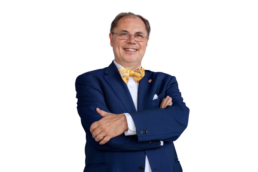 Un uomo sorridente in giacca blu e cravatta a farfalla colorata, con le braccia incrociate, su sfondo bianco.