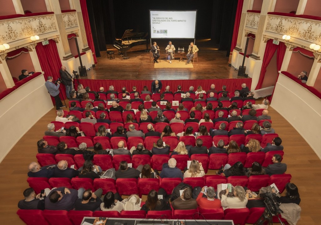 Cerimonia di premiazione al Teatro degli Illuminati di Città di Castello, con una folla di spettatori seduti in poltrone rosse, mentre i relatori sono sul palco.
