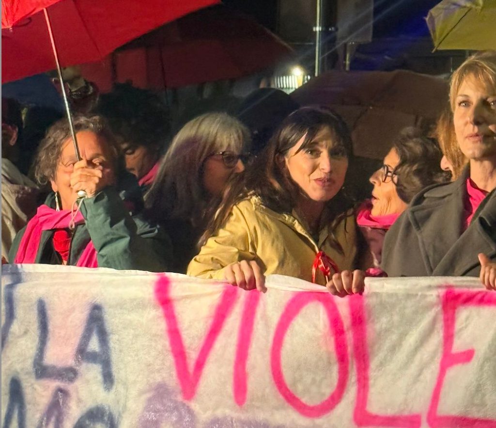 Un gruppo di donne partecipa a una manifestazione contro la violenza maschile, tenendo uno striscione con scritte in evidenza. Alcune donne indossano sciarpe rosa e ci sono ombrelli aperti sullo sfondo.