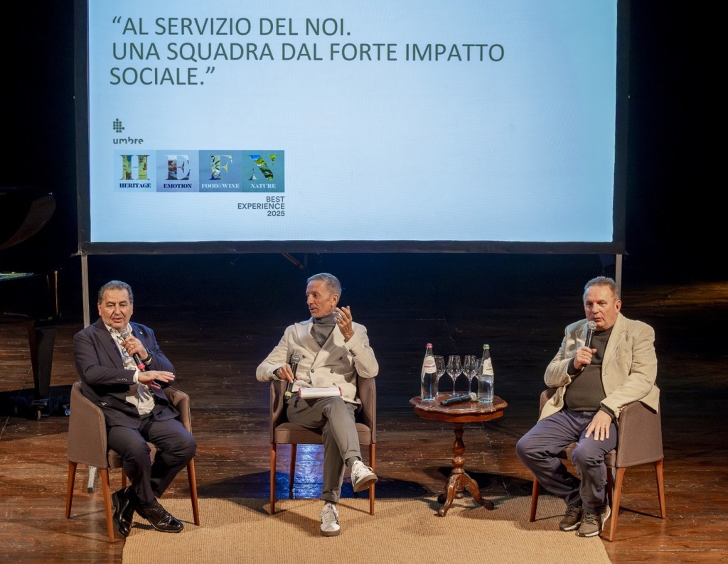 Tre relatori discutono su un palco mentre un proiettore mostra il messaggio 'AL SERVIZIO DEL NOI. UNA SQUADRA DAL FORTE IMPATTO SOCIALE.' durante un evento per il Premio 'Best Experience 2025' a Città di Castello.