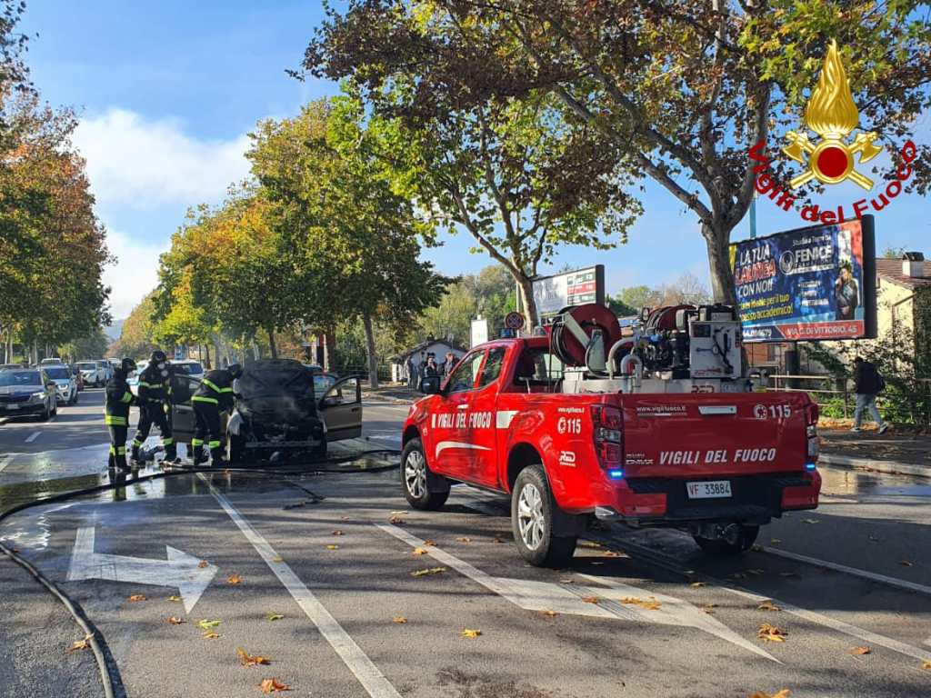 Firefighters responding to a car fire on a street surrounded by trees and parked cars.