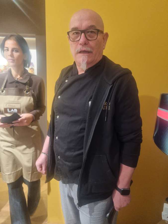 Un maestro pasticcere in un laboratorio di cioccolato, con una donna di spalle che indossa un grembiule, davanti a uno sfondo colorato.