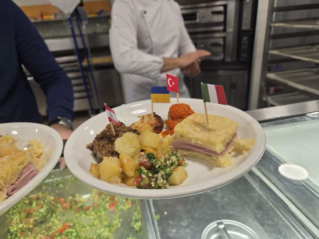 Piatto di cibo con diverse preparazioni, incluse bandierine di Ucraina, Turchia e Italia, presentato al concorso gastronomico 'Da leccarsi i baffi'.