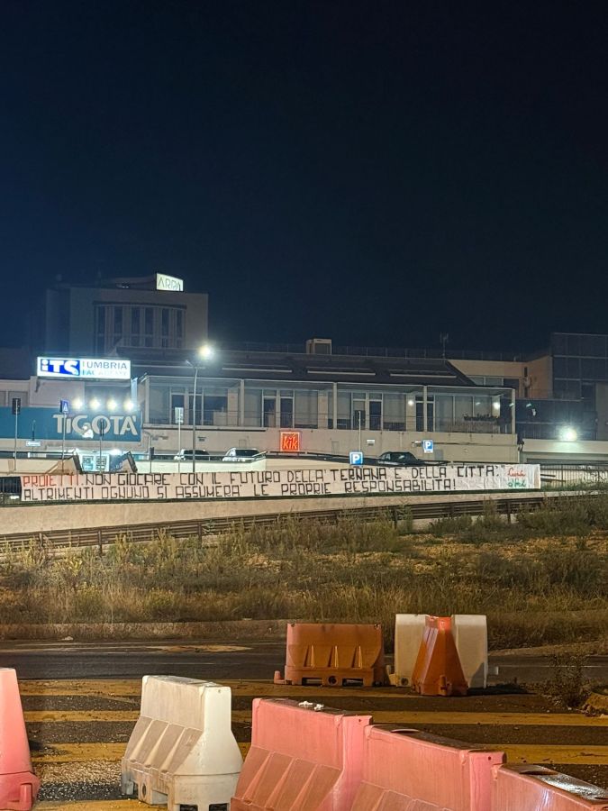 Sign on a building at night urging citizens to take responsibility for the future of Terni and its football club, with construction barriers in the foreground.