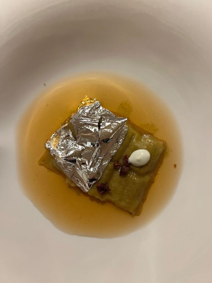 Un piatto gourmet con un raviolo avvolto in foglio d'alluminio, servito su un letto di salsa dorata e guarnito con crema bianca e petali di fiori.