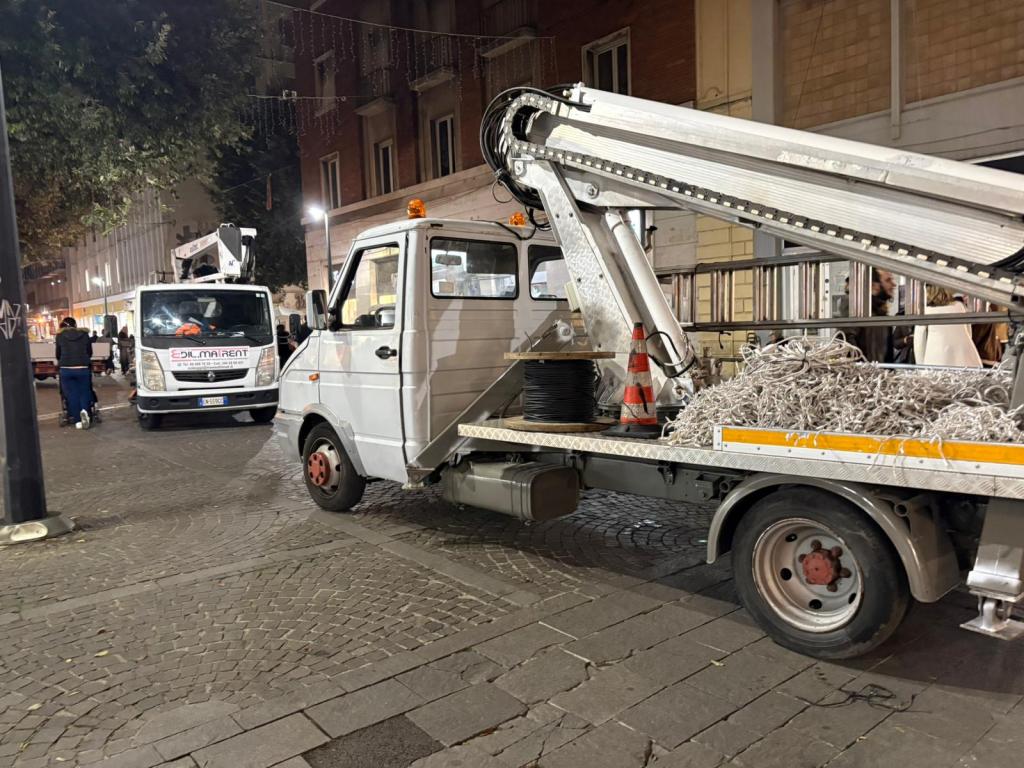 Un furgón blanco estacionado en una calle, con un brazo mecánico extendido y cables en la plataforma trasera, mientras otro vehículo de trabajo se encuentra al fondo en un entorno urbano nocturno.