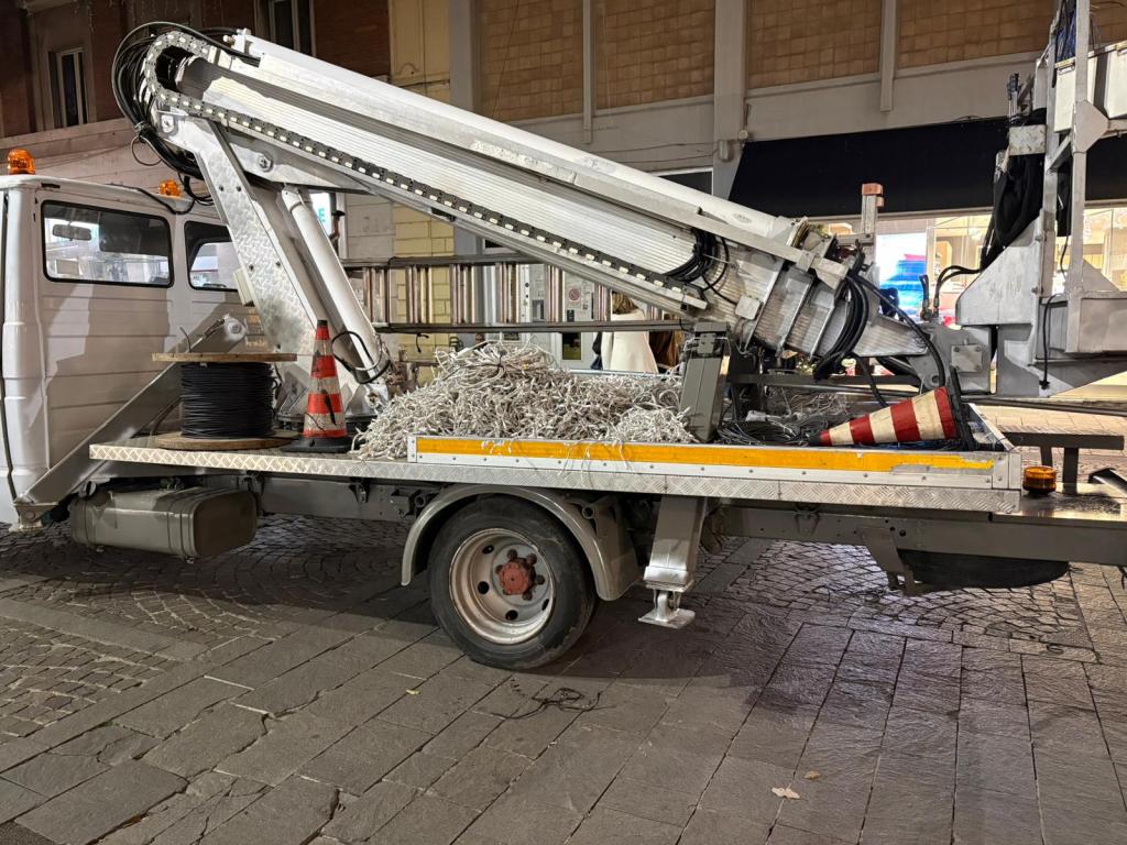 A utility truck parked on a cobblestone street, featuring an extendable arm and a spool of wire, with a pile of ropes and a traffic cone on the flatbed.