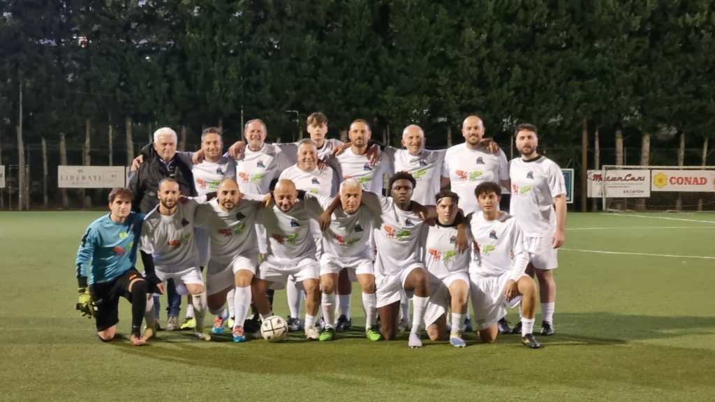 Foto di un gruppo di giocatori di calcio in magliette bianche, sorridenti e in posa, sul campo da gioco, con alberi sullo sfondo.