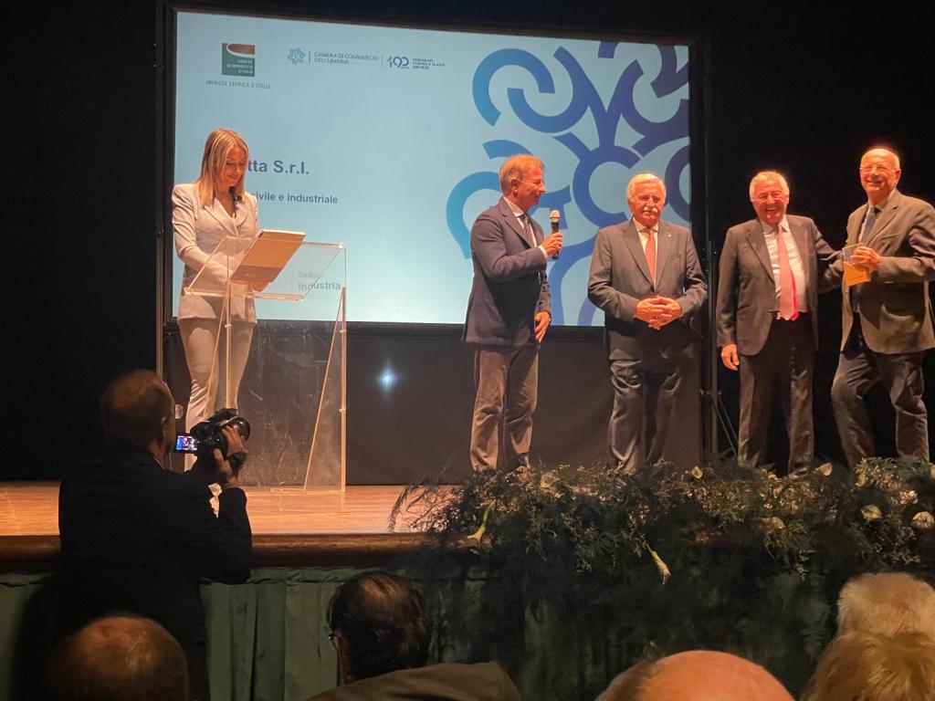 Cerimonia di premiazione con un presentatore e diversi relatori sul palco, mentre il pubblico osserva. Un ambiente elegante con decorazioni floreali.
