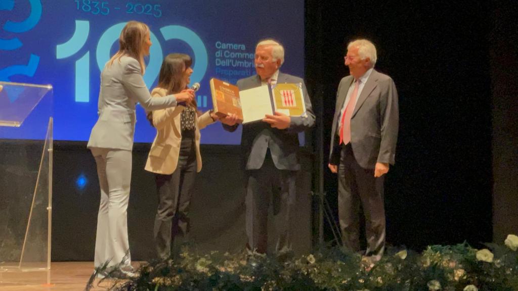 Cerimonia di premiazione con tre persone che ricevono riconoscimenti sul palco, tra cui un uomo con un gesto di orgoglio mentre mostra un premio.