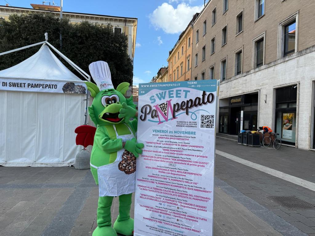 Un personaggio mascotte sorridente in costume verde con adorni culinari, mentre tiene un poster pubblicitario per l'evento Sweet Pampepato in Piazza Tacito a Terni.