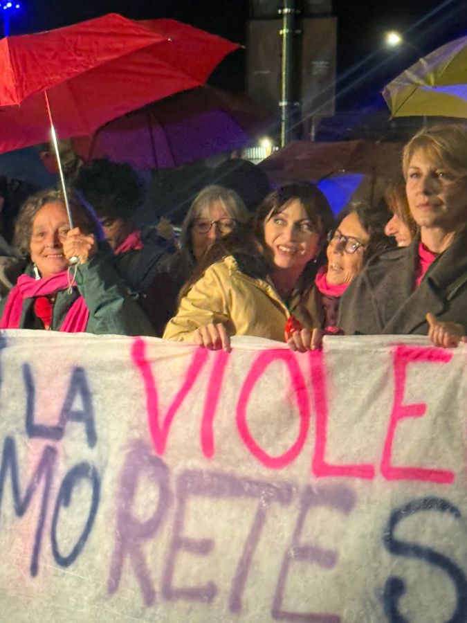 Un gruppo di donne partecipa a una manifestazione contro la violenza di genere, tenendo in mano un grande striscione. Alcune donne utilizzano ombrelli per ripararsi dalla pioggia.