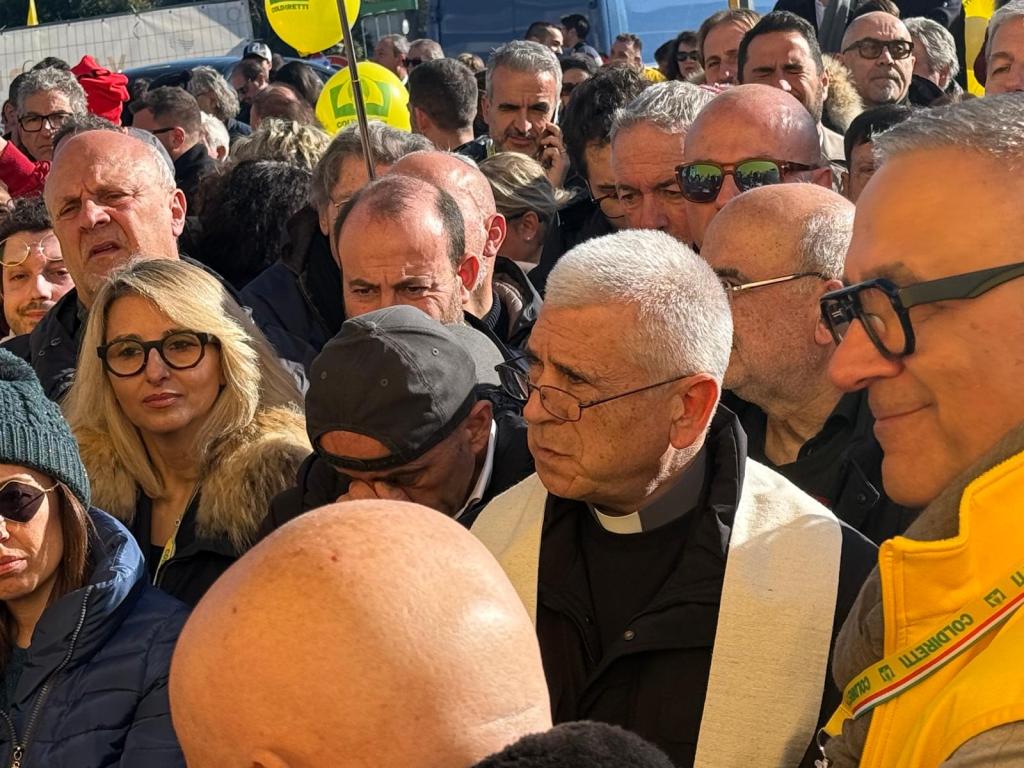 A large crowd of people is gathered, with diverse expressions and attire, highlighting a moment of communal focus. A person in clerical attire stands prominently among the crowd.