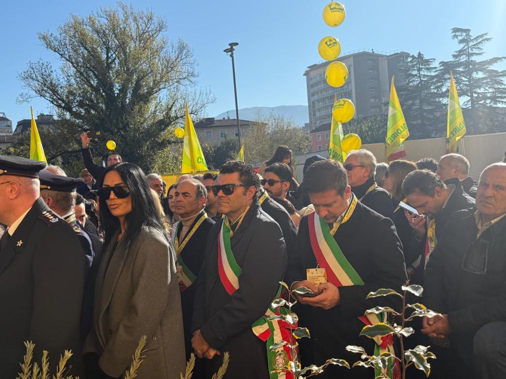 A crowd gathered outdoors, with individuals wearing sashes in the colors of the Italian flag and holding yellow flags. A woman in sunglasses stands prominently in the foreground, while others appear engaged in their phones.