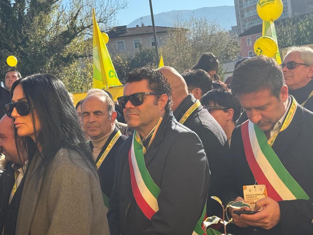 A group of people in formal attire, some wearing sashes with the Italian flag colors, gathered outdoors during a sunny event with banners and flags in the background.