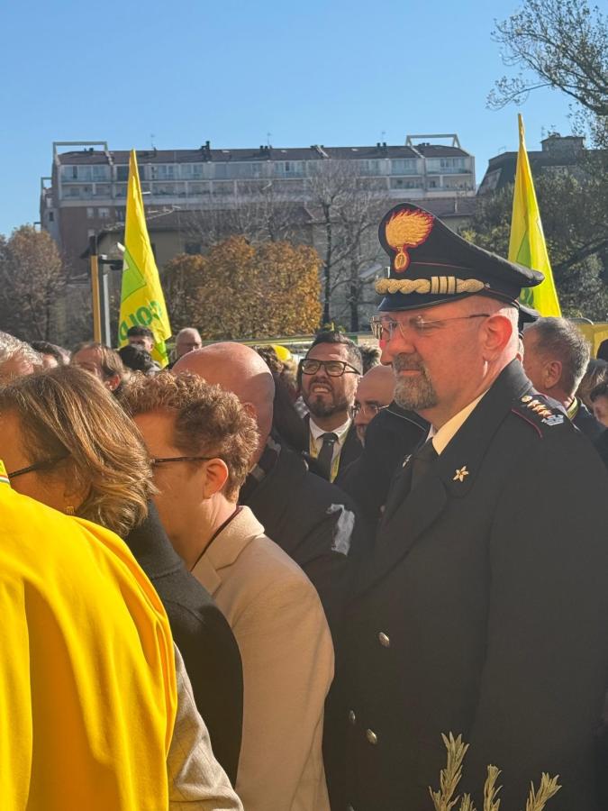 A crowd of people outdoors, including a man in a police uniform with a cap, surrounded by others wearing suits and yellow banners in the background.