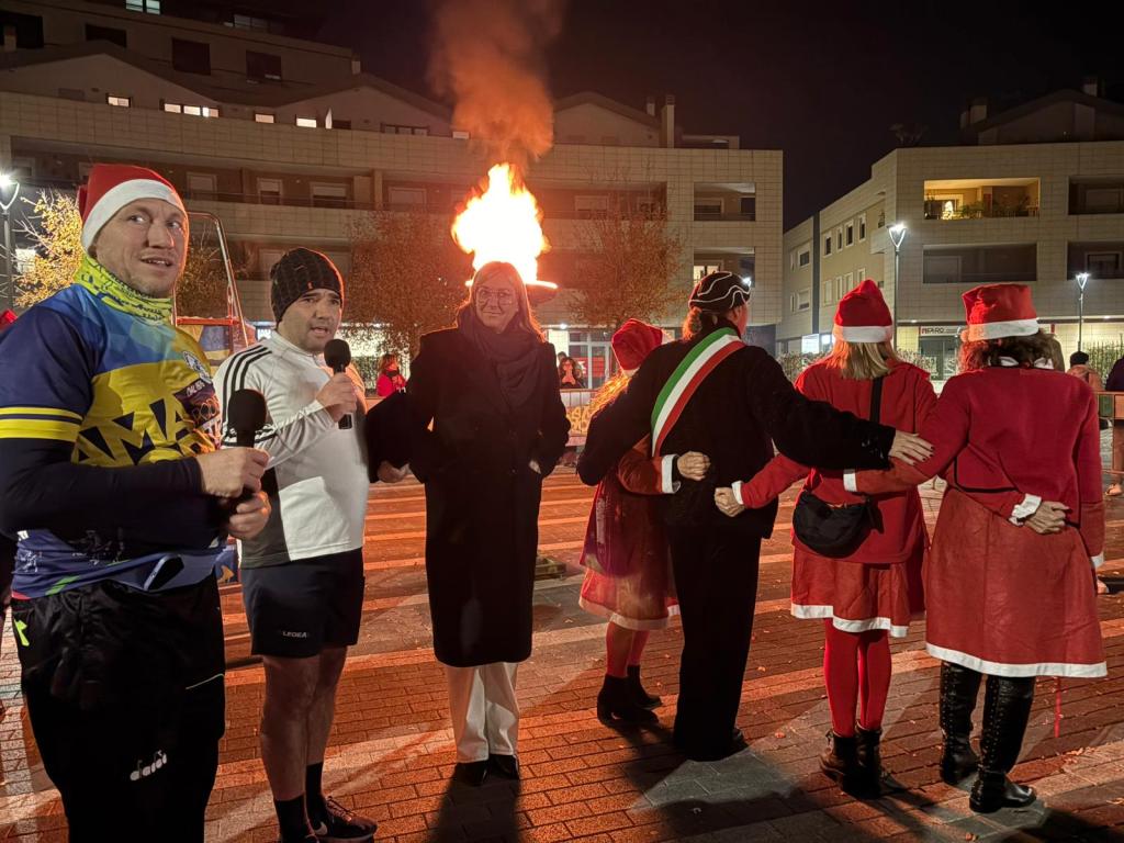 A group of people gathered outdoors at night, with two men speaking into microphones and a fire burning in the background. Several individuals are dressed in festive Christmas attire, including Santa hats and red costumes, while others appear to be wearing casual clothing.