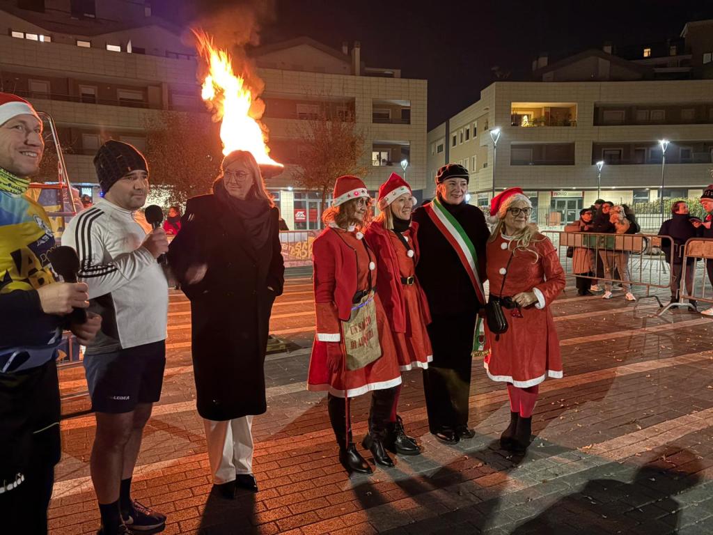 A group of people celebrating outdoors at night, with a fire in the background. Some participants are dressed in festive attire, including Santa hats and red dresses. A man in sportswear is speaking into a microphone, while a woman in a black coat looks on.