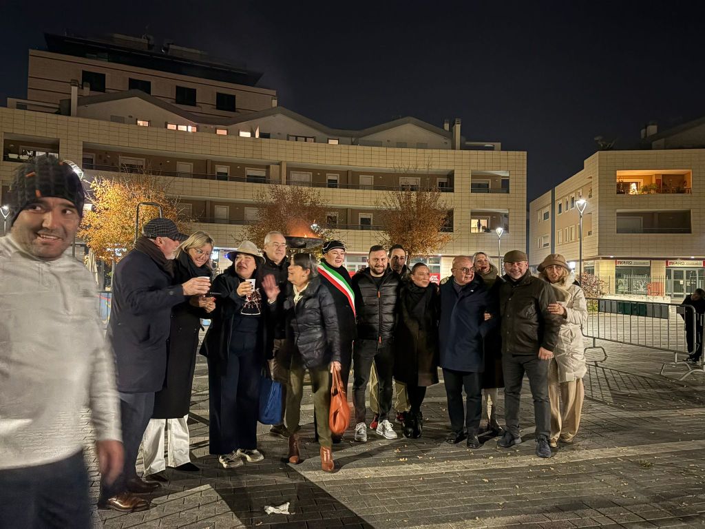 A group of people celebrating outdoors at night, surrounded by modern buildings, with some holding drinks and smiling.