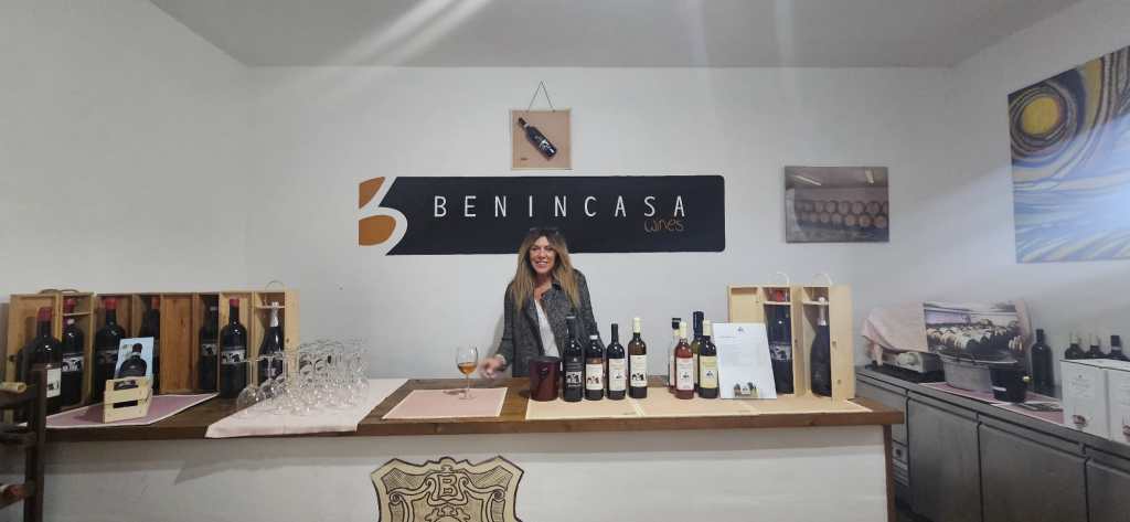 Una donna sorride dietro il bancone di una cantina vinicola, con bottiglie di vino e bicchieri visibili sul tavolo e un'insegna con il nome 'Benincasa' sullo sfondo.