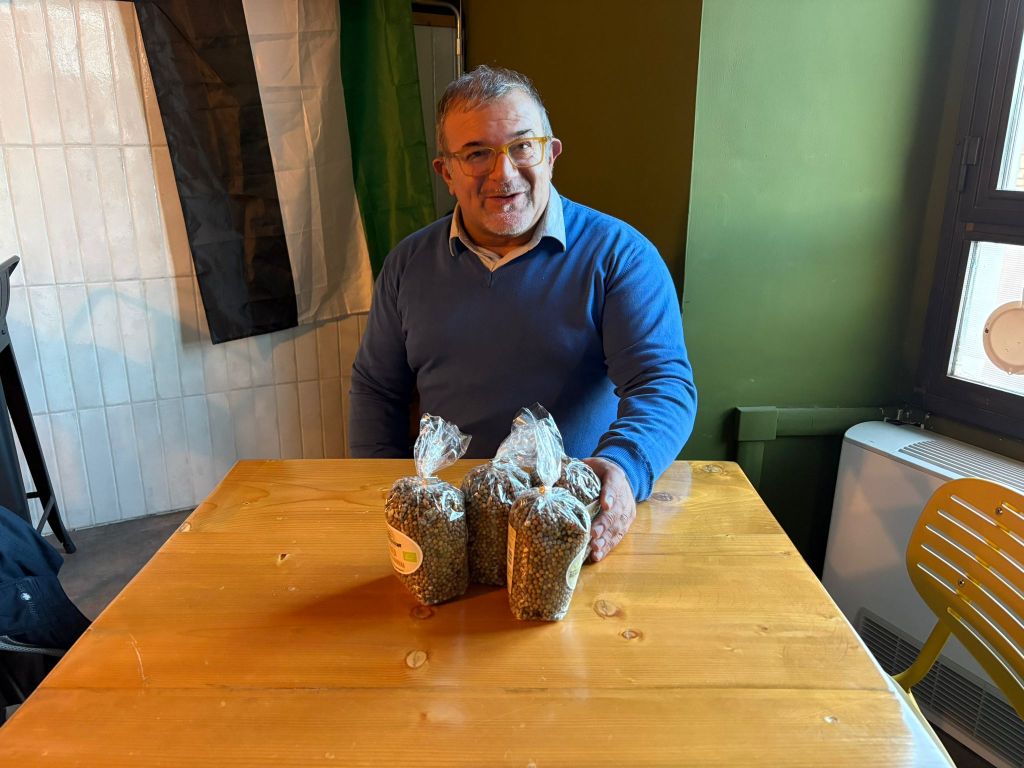 Un uomo sorridente seduto a un tavolo di legno, con in testa una maglietta blu, che tiene tre sacchetti di roveja, un legume antico, mentre si trova accanto a una bandiera e una parete verde.
