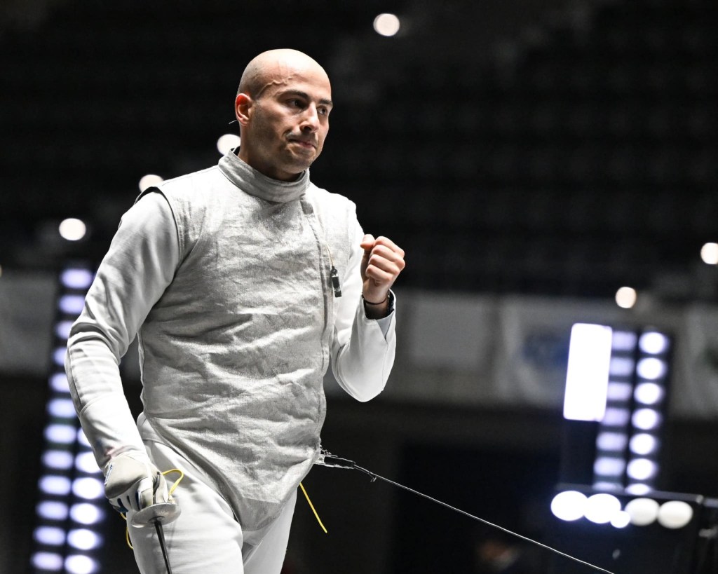 Un schermidore con un'espressione determinata mentre solleva il pugno in segno di vittoria, indossando un'uniforme da scherma grigia in un ambiente sportivo.