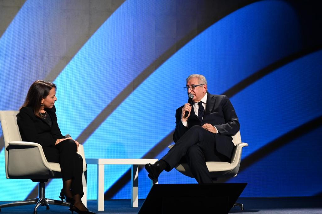 Due relatori seduti su poltrone bianche durante un evento, con uno dei relatori che parla al microfono. Sullo sfondo, un'illuminazione blu.