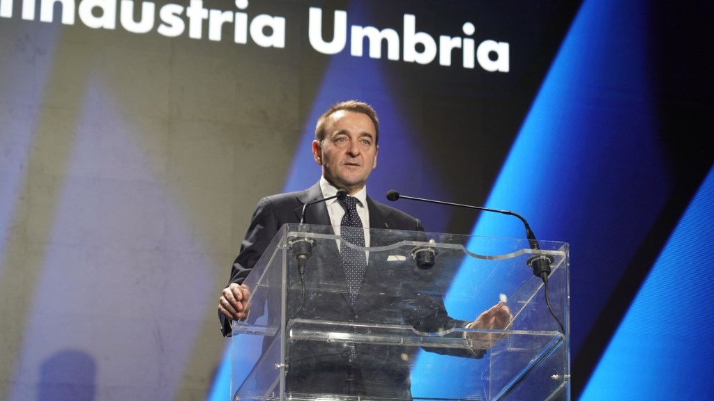 Un uomo in abito formale parla al microfono davanti a un podio trasparente, con uno sfondo che presenta il logo di Confindustria Umbria.