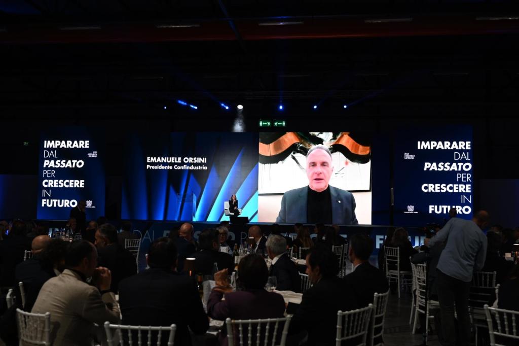 Evento di gala per l'Assemblea pubblica di Confindustria Umbria 2025, con un relatore proiettato su uno schermo e pubblico seduto a tavoli.