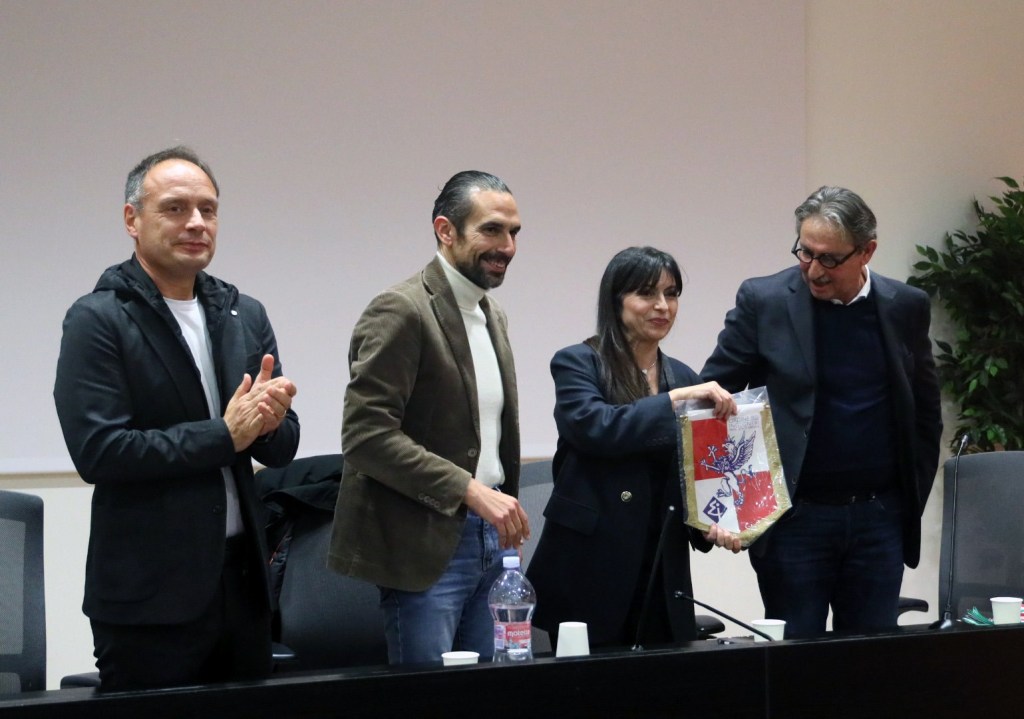 Un gruppo di persone al tavolo di una conferenza, con un uomo a sinistra che applaude, un altro uomo al centro con un sorriso, una donna che tiene un premio e un uomo a destra che sorride mentre osserva.